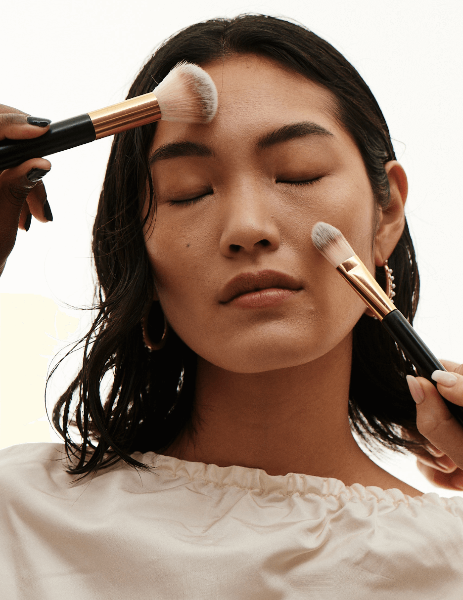 Female model posing while two other hands are using makeup brushes on her face. 